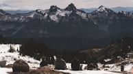 A group of people sitting on a snowy hill with backpacks, overlooking a vast mountainous landscape. The peaks in the background are covered with patches of snow and surrounded by dense forests.