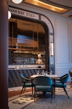 A sophisticated restaurant interior featuring a raw bar. The setting is elegant with mosaic tiles on the wall displaying 'BRASSERIE 1783' and 'RAW BAR'. The dining area includes teal velvet chairs and a neatly set table. A chef is visible through a window into the kitchen, wearing a traditional white chef's hat.