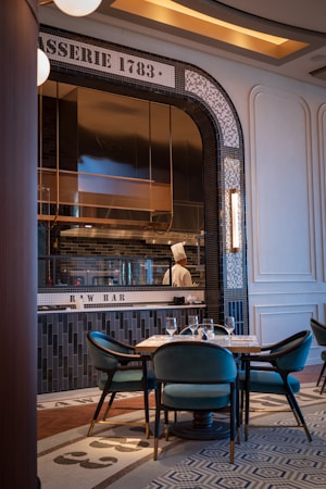A sophisticated restaurant interior featuring a raw bar. The setting is elegant with mosaic tiles on the wall displaying 'BRASSERIE 1783' and 'RAW BAR'. The dining area includes teal velvet chairs and a neatly set table. A chef is visible through a window into the kitchen, wearing a traditional white chef's hat.