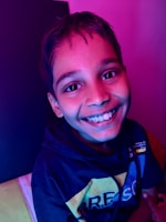 A young person smiling broadly with vibrant lighting casting shades of pink and blue on their face. They are wearing a dark hoodie with colorful text across the front.