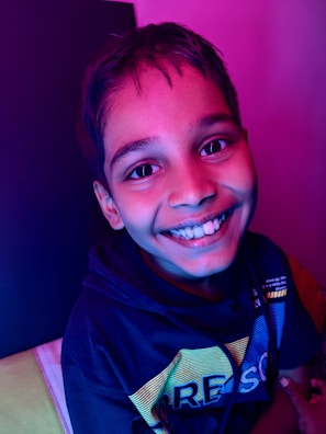 A young person smiling broadly with vibrant lighting casting shades of pink and blue on their face. They are wearing a dark hoodie with colorful text across the front.