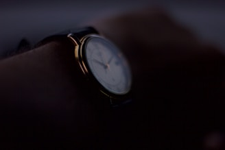 A close-up of a wristwatch on a person's wrist, with a dimly lit setting that creates a mysterious and subdued atmosphere. The watch face is partially visible, with metallic elements catching faint light.