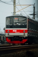 A KAI Commuter train approaches on a railway track against a backdrop of overhead electric lines and a clear sky. The train is primarily red and grey, with visible text indicating its route to Bekasi.