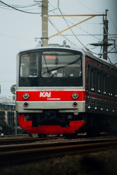A KAI Commuter train approaches on a railway track against a backdrop of overhead electric lines and a clear sky. The train is primarily red and grey, with visible text indicating its route to Bekasi.