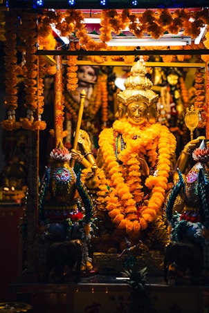 A golden deity statue adorned with vibrant orange marigold garlands is surrounded by elaborately decorated elements, including ornate carvings and small elephant statues. The setting is richly detailed and festive with bright marigold flowers draped throughout.