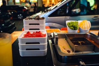 A table is set for hot pot dining with a metal pot divided into sections containing different broths. Nearby are stacks of white trays with raw meat and seafood. On the table, there are also bowls filled with fresh green lettuce and neatly arranged taco shells. In the background, a busy street with cars and people creates a lively urban atmosphere.