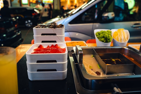 A table is set for hot pot dining with a metal pot divided into sections containing different broths. Nearby are stacks of white trays with raw meat and seafood. On the table, there are also bowls filled with fresh green lettuce and neatly arranged taco shells. In the background, a busy street with cars and people creates a lively urban atmosphere.