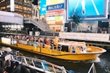 A vibrant yellow tourist boat is navigating through a canal in an urban area at night. The boat is filled with passengers who are observing the bustling environment. Brightly lit billboards and signs, along with tall buildings, surround the canal, contributing to an energetic atmosphere. Pedestrians can be seen on footbridges and docks nearby.