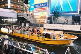 A vibrant yellow tourist boat is navigating through a canal in an urban area at night. The boat is filled with passengers who are observing the bustling environment. Brightly lit billboards and signs, along with tall buildings, surround the canal, contributing to an energetic atmosphere. Pedestrians can be seen on footbridges and docks nearby.