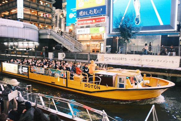 A vibrant yellow tourist boat is navigating through a canal in an urban area at night. The boat is filled with passengers who are observing the bustling environment. Brightly lit billboards and signs, along with tall buildings, surround the canal, contributing to an energetic atmosphere. Pedestrians can be seen on footbridges and docks nearby.