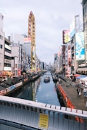 A vibrant urban scene features a bustling canal flanked by tall buildings adorned with colorful advertisements. A Ferris wheel rises prominently, adding to the lively cityscape. Pedestrians walk along the canal, while a tourist boat glides through the water.