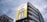 A large advertisement on the side of a building displays a can of Asahi Super Dry beer with a vibrant yellow background. The building is multi-storied with modern architecture, and the sky is overcast with grayish clouds.