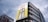 A large advertisement on the side of a building displays a can of Asahi Super Dry beer with a vibrant yellow background. The building is multi-storied with modern architecture, and the sky is overcast with grayish clouds.