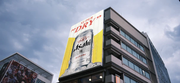 A large advertisement on the side of a building displays a can of Asahi Super Dry beer with a vibrant yellow background. The building is multi-storied with modern architecture, and the sky is overcast with grayish clouds.