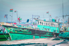 A green and black boat is docked at a busy harbor. The boat is accompanied by several other vessels, and there are Indian flags flying from some masts. The scene captures a vibrant marine setting with ropes, nets, and equipment scattered around the dock.