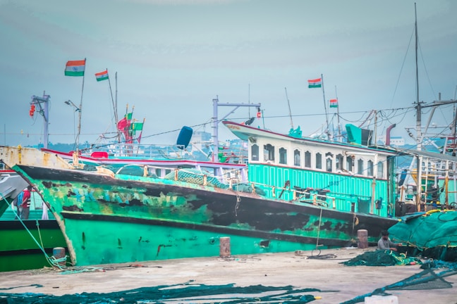 A green and black boat is docked at a busy harbor. The boat is accompanied by several other vessels, and there are Indian flags flying from some masts. The scene captures a vibrant marine setting with ropes, nets, and equipment scattered around the dock.