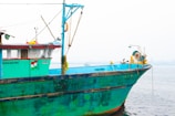 A colorful wooden fishing boat is docked by the water, featuring bright turquoise and green paint. The boat is equipped with various fishing gear, ropes, and a winch system. An Indian flag is painted on the side, and the registration number is visible. The background shows calm water with a slight haze obscuring the distant horizon.