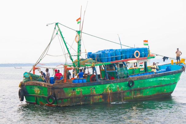 A fishing boat is stationed in the water, adorned with flags and numerous blue barrels on top. Several people are visible on the boat, some engaging in work-related activities. The vessel is painted green with visible wear and signs of rust, indicating its age and usage.