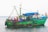 A fishing boat is stationed in the water, adorned with flags and numerous blue barrels on top. Several people are visible on the boat, some engaging in work-related activities. The vessel is painted green with visible wear and signs of rust, indicating its age and usage.