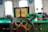 A ship's wheel is centered in the image, made of wood and metal. The background features a green dashboard with multiple windows letting in natural light. Various equipment and controls, including levers and a flashlight, are visible. A box labeled 'SHIHANG' is situated behind the wheel.