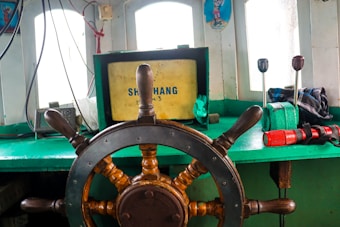 A ship's wheel is centered in the image, made of wood and metal. The background features a green dashboard with multiple windows letting in natural light. Various equipment and controls, including levers and a flashlight, are visible. A box labeled 'SHIHANG' is situated behind the wheel.