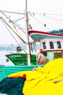 A fishing boat with a red and white cabin sits docked next to another green vessel named 'St. Peter.' A person wearing a bright pink headscarf and a weathered, beige jacket stands on the deck near large bundles of yellow fabric and fishing nets. The background features a hazy waterfront view with buildings and trees.