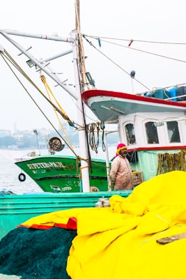 A fishing boat with a red and white cabin sits docked next to another green vessel named 'St. Peter.' A person wearing a bright pink headscarf and a weathered, beige jacket stands on the deck near large bundles of yellow fabric and fishing nets. The background features a hazy waterfront view with buildings and trees.