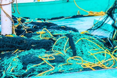 A tangled jumble of brightly colored fishing nets in shades of teal and yellow, resting on a boat deck. Some ropes are intertwined with the nets, creating an intricate and chaotic pattern. A white post is visible, possibly part of the boat structure.
