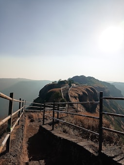 A pathway with metal railings edges along a cliff with expansive views of distant hills and a bright sun in the sky. The ground has dry grass, and the path leads to a higher elevation with sparse vegetation.