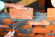 A pair of hands are working on building a brick wall. One hand is placing an orange brick onto a layer of wet gray mortar, while the other hand is holding a trowel, which is likely being used to spread the mortar. The bricks are arranged in a staggered pattern, and a taut string line is used as a guide for alignment.