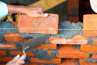 A pair of hands are working on building a brick wall. One hand is placing an orange brick onto a layer of wet gray mortar, while the other hand is holding a trowel, which is likely being used to spread the mortar. The bricks are arranged in a staggered pattern, and a taut string line is used as a guide for alignment.