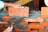 A pair of hands are working on building a brick wall. One hand is placing an orange brick onto a layer of wet gray mortar, while the other hand is holding a trowel, which is likely being used to spread the mortar. The bricks are arranged in a staggered pattern, and a taut string line is used as a guide for alignment.