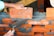 A pair of hands are working on building a brick wall. One hand is placing an orange brick onto a layer of wet gray mortar, while the other hand is holding a trowel, which is likely being used to spread the mortar. The bricks are arranged in a staggered pattern, and a taut string line is used as a guide for alignment.
