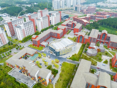 An aerial view of a large urban complex featuring multiple modern buildings with red and white facades surrounded by greenery. The layout includes several rectangular structures with flat roofs, wide courtyards, and open spaces. In the distance, there are taller residential buildings with consistent architectural styles.
