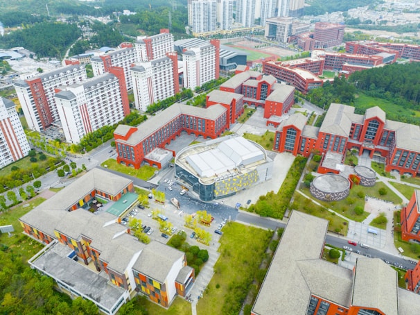 An aerial view of a large urban complex featuring multiple modern buildings with red and white facades surrounded by greenery. The layout includes several rectangular structures with flat roofs, wide courtyards, and open spaces. In the distance, there are taller residential buildings with consistent architectural styles.
