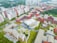 An aerial view of a large urban complex featuring multiple modern buildings with red and white facades surrounded by greenery. The layout includes several rectangular structures with flat roofs, wide courtyards, and open spaces. In the distance, there are taller residential buildings with consistent architectural styles.