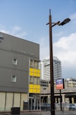 A modern urban scene featuring a tall brown streetlamp in the foreground and a building with a grey facade and yellow paneling in the background. There are additional buildings and a highway in the distance, conveying a cityscape environment. Visible signage includes some branding.