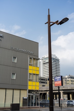 A modern urban scene featuring a tall brown streetlamp in the foreground and a building with a grey facade and yellow paneling in the background. There are additional buildings and a highway in the distance, conveying a cityscape environment. Visible signage includes some branding.