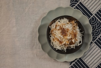 A bowl filled with shredded white coconut topped with caramel or brown syrup sits on a dark plate with a scalloped edge. The setup is placed on a beige fabric surface partially covering a patterned dark and light cloth.