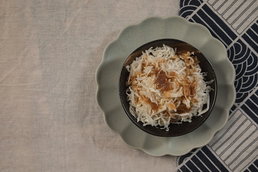 A bowl filled with shredded white coconut topped with caramel or brown syrup sits on a dark plate with a scalloped edge. The setup is placed on a beige fabric surface partially covering a patterned dark and light cloth.