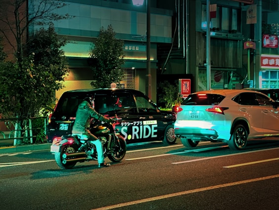 A nighttime street scene featuring a motorcyclist in a helmet and jacket stopped beside a black taxi with 'S.RIDE' written on its side and a white SUV in front. The street is lined with buildings and trees, illuminated by neon lights with a greenish hue creating reflections on the vehicles.