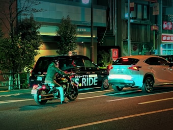 A nighttime street scene featuring a motorcyclist in a helmet and jacket stopped beside a black taxi with 'S.RIDE' written on its side and a white SUV in front. The street is lined with buildings and trees, illuminated by neon lights with a greenish hue creating reflections on the vehicles.