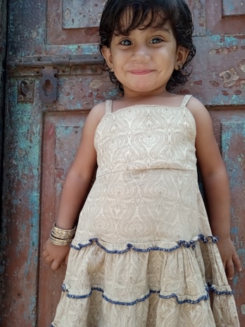 A young child wearing a beige dress with blue trim stands against a weathered metal door, smiling warmly. The child has short, dark hair and is adorned with bangles on one wrist.
