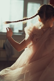 A young girl twirls in a softly lit room, her dress flowing around her in delicate layers of fabric. Sunlight streams through a window, highlighting her braided hair and creating a dreamy, ethereal atmosphere.
