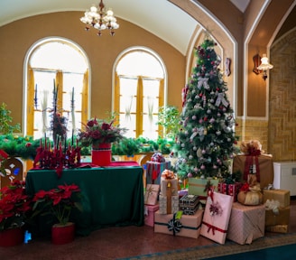 A festive indoor setting featuring a decorated Christmas tree adorned with ornaments and ribbons, situated next to a pile of wrapped gifts. There is a table covered with a green cloth that holds a red centerpiece, candles, and various Christmas decorations. Red poinsettias and green garlands add to the holiday atmosphere, with arched windows in the background letting in natural light.
