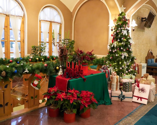 A decorated room featuring a Christmas tree adorned with lights, bows, and ornaments. Presents wrapped in festive paper are placed around the tree. A green and red-themed table is set with red candles and floral arrangements. Poinsettias in pots and green garlands with ornaments enhance the holiday decor. Three arched windows with curtains add a cozy ambiance.