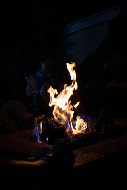 A fire pit burns brightly in the center with orange and blue flames. The area is dimly lit, suggesting a nighttime setting. Three people are seated around the fire, partially visible due to the darkness. They appear relaxed and engaged, enjoying the warmth of the fire.