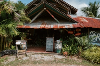 A rustic, tropical-themed building with a corrugated metal roof and wooden structure. The entrance is flanked with lush green plants, and there are signboards displaying the menu and the name of the establishment. The building appears to be a casual dining or cafe setup, with seating visible inside.