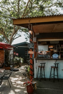 A small outdoor café setup with a rustic charm. The scene includes a counter with stools, surrounded by greenery and decorative plants in pots. There's a sign indicating that the café is open and some glassware on display. The area is shaded by trees and a corrugated metal roof.