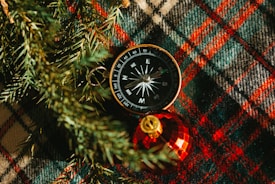 A compass is placed on a plaid fabric with shades of green, red, and brown. To the bottom right of the compass is a shiny red Christmas ornament with a golden top. Pine needles are visible around the compass, adding a festive touch.